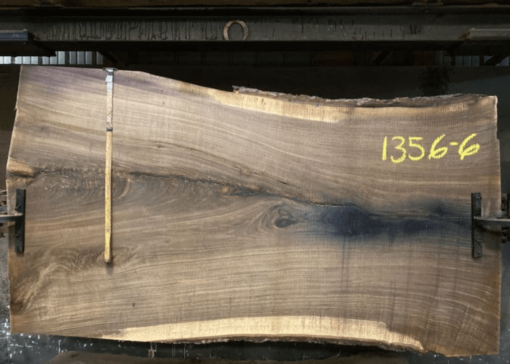 A large, polished Walnut slab with natural edges is secured in a horizontal position. The slab showcases a mix of brown shades and a unique grain pattern. A wooden ruler is placed vertically for scale, and the numbers "1356-6" are written in yellow on the upper right corner.